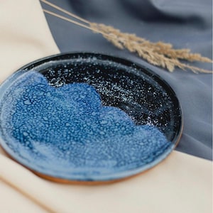 May include: A handmade ceramic plate with a unique design. The plate features a dark blue and black glazed surface with a speckled pattern, and a lighter blue section. The rim has a natural, unglazed appearance.