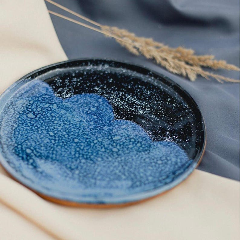 May include: A handmade ceramic plate with a unique design. The plate features a dark blue and black glazed surface with a speckled pattern, and a lighter blue section. The rim has a natural, unglazed appearance.