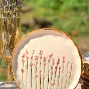 May include: A handmade ceramic plate with a lavender design. The plate has a light beige background with a brown rim and a wooden stand. The lavender stems are pink and green. A vase of lavender is in the background.