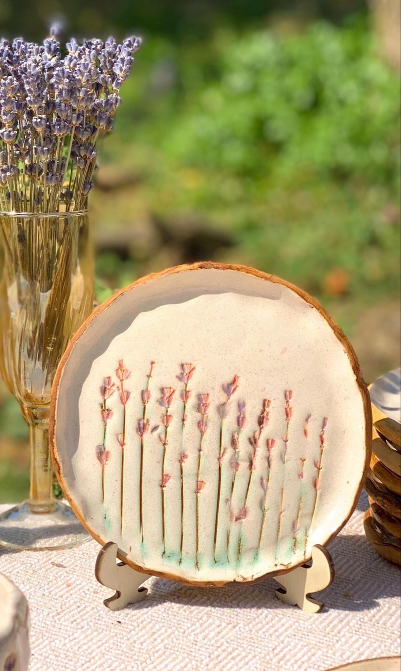 May include: A handmade ceramic plate with a lavender design. The plate has a light beige background with a brown rim and a wooden stand. The lavender stems are pink and green. A vase of lavender is in the background.