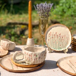 May include: Handmade ceramic teacup and saucer set with a floral design. The set includes a teacup, saucer, and small bowl, all with a textured, off-white finish and hand-painted pink and green floral accents. A candle and lavender bouquet are also in the image.