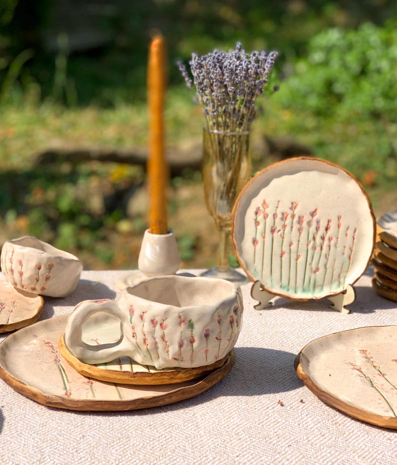 May include: Handmade ceramic teacup and saucer set with a floral design. The set includes a teacup, saucer, and small bowl, all with a textured, off-white finish and hand-painted pink and green floral accents. A candle and lavender bouquet are also in the image.