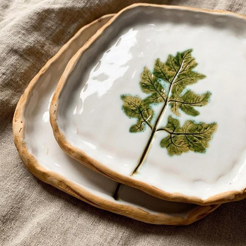 May include: Two white ceramic plates with a leaf design. The plates have a rustic, irregular edge and a brown rim. The leaf is green and brown, with a detailed vein pattern. The plates are stacked on top of each other.