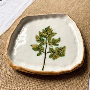 May include: A handmade ceramic plate with a white glazed surface and a brown, organic edge. The plate features a detailed green leaf design in the centre. The plate is approximately 20 cm wide.