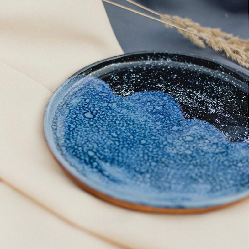May include: A ceramic plate with a unique design. The plate features a dark blue and black speckled rim, with a wave-like pattern of textured blue glaze. The plate rests on a cream-colored surface, with a sprig of dried grass in the background.