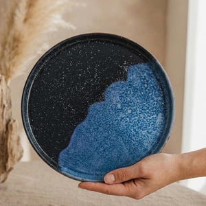 May include: A round ceramic plate with a two-tone design. The top half is black with white speckles, and the bottom half is a textured blue. The plate is held in a person's hand.