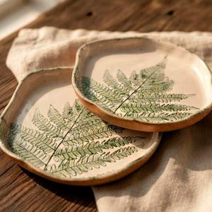 May include: Two handmade ceramic plates with irregular, organic shapes. Each plate features a cream-coloured glaze and a detailed, hand-painted green fern leaf design that spans across the surface, showcasing natural textures and earthy tones.
