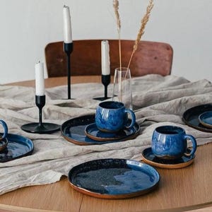 May include: A table setting featuring blue ceramic mugs and plates with a speckled glaze. The set includes matching saucers. Black candle holders with white candles and a vase with dried plants complete the arrangement.