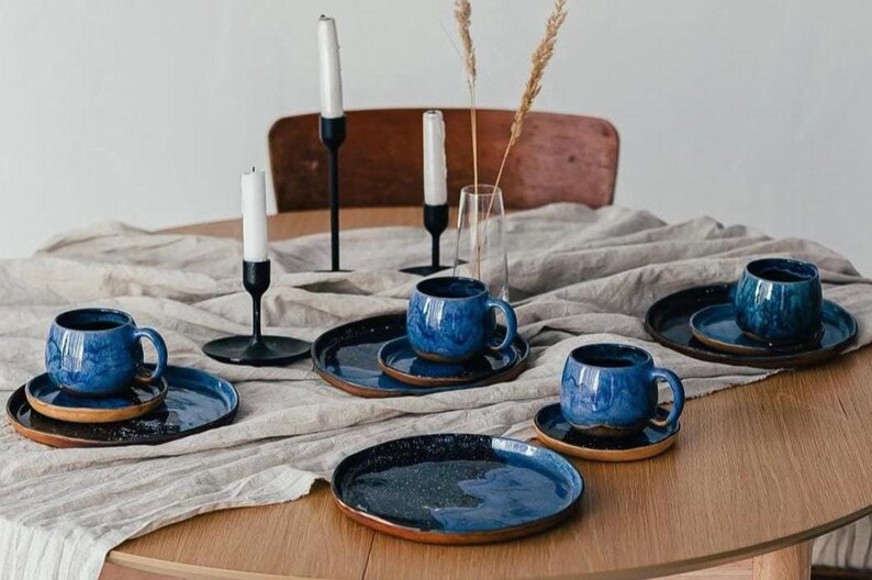 May include: A table setting featuring blue ceramic mugs and plates with a speckled glaze. The set includes matching saucers. Black candle holders with white candles and a vase with dried plants complete the arrangement.