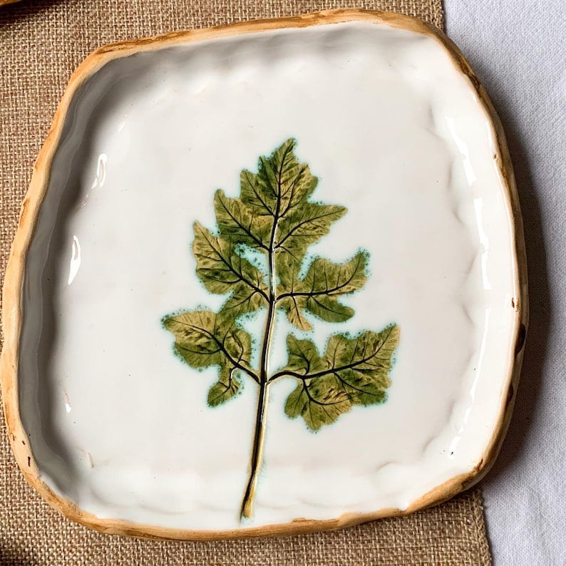 May include: Square ceramic plate with a white surface and a raised, irregular edge. The plate features a detailed green and brown leaf design in the centre. The rim has a natural, earthy tone.