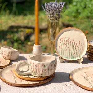 May include: Handmade ceramic teacup and saucer set with a floral design. The set includes a teacup, saucer, and small plate, all with a cream-colored base and delicate pink and green floral accents. A small bowl and a decorative plate are also visible.