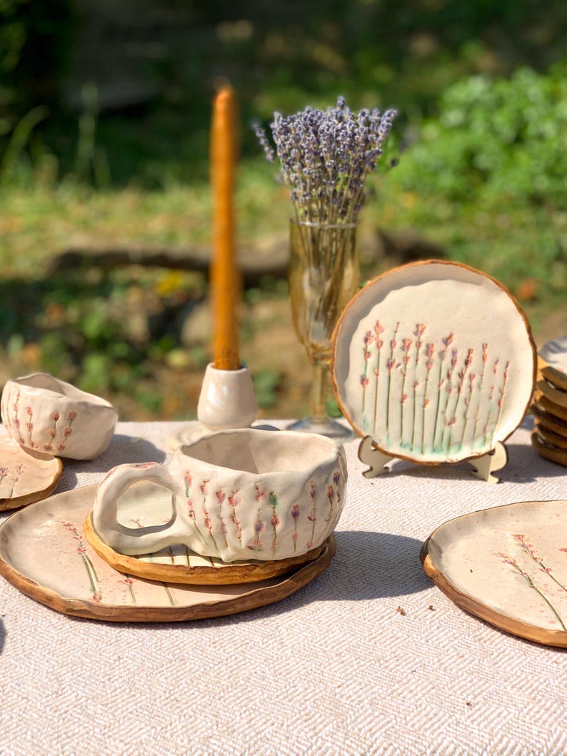 May include: Handmade ceramic teacup and saucer set with a floral design. The set includes a teacup, saucer, and small plate, all with a cream-colored base and delicate pink and green floral accents. A small bowl and a decorative plate are also visible.