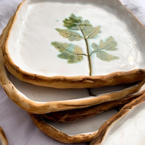 May include: A stack of handmade ceramic plates. Each plate has a white surface with a green and gold leaf design. The edges are a natural brown colour, giving them a rustic appearance. The plates are stacked on top of each other.