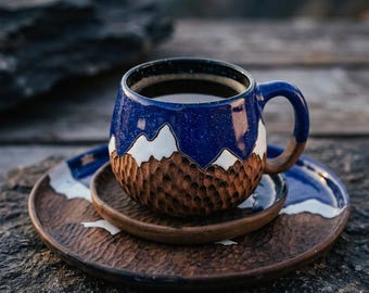Może przedstawiać: Zestaw ceramiczny składający się z kubka i spodka, z ciemnoniebieskim nakrapianym szkliwem w górnej części oraz rzeźbioną, brązową, teksturowaną podstawą. Wzór zawiera białe szczyty górskie oddzielające niebieskie i brązowe sekcje na obu elementach.
