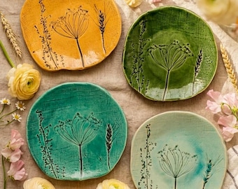 May include: Four round ceramic plates in yellow, dark green, teal, and light blue, each featuring a textured grid surface and a black botanical illustration of a wildflower and wheat stalk. The plates have irregular, organic edges and smooth finishes.