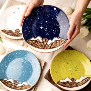 Op de afbeelding: Een set van vier keramische borden met een bergontwerp. De borden hebben een gespikkelde afwerking in blauw, wit en geel, met een donkerblauw bord in het midden. Elk bord heeft een bergketenontwerp aan de onderkant.