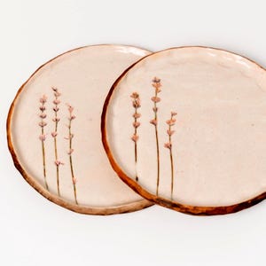 Op de afbeelding: Twee ronde keramische borden met een gebroken wit, gespikkeld oppervlak en onregelmatige, donkerbruine randen. Elk bord is voorzien van een delicate, handgeschilderde botanische illustratie van drie slanke lavendelstengels met kleine, lichtpaarse bloemknoppen.