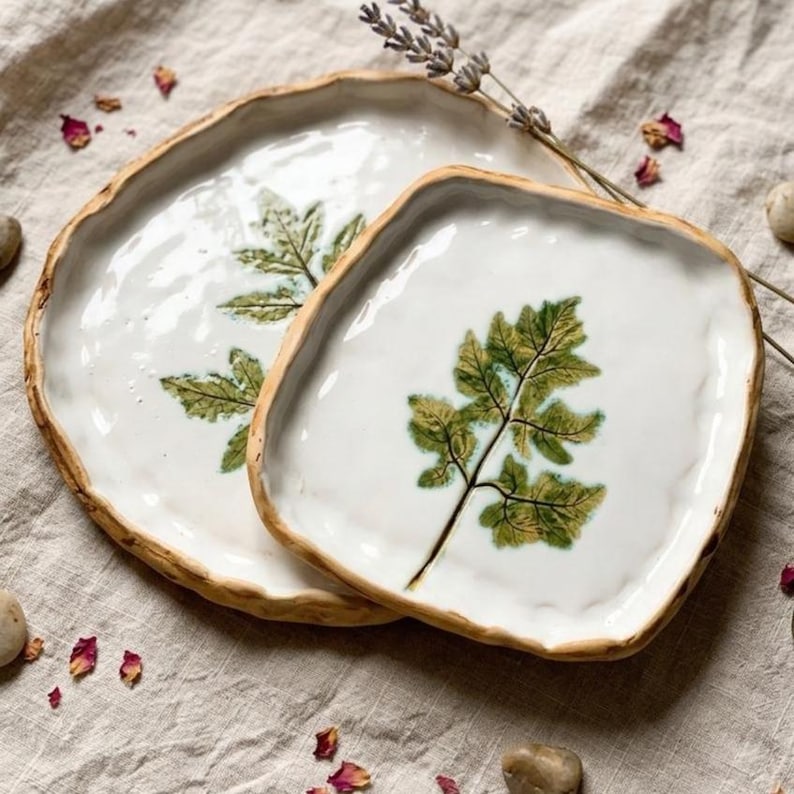 May include: Two white ceramic plates with a leaf design. The plates have a natural, organic shape with a rough, brown edge. The leaf is green and brown, and the plates are on a neutral-coloured surface.