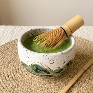 Può includere: Una ciotola di matcha in ceramica con tè verde e un frullino di bambù. La ciotola ha una base bianca con macchie blu e un disegno paesaggistico in verde e blu. Un cucchiaio di bambù è appoggiato di lato.