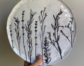 May include: A round, off-white ceramic plate with a speckled finish and an uneven, organic edge. The surface features dark blue, embossed botanical imprints of various wildflowers and stems arranged vertically across the center of the plate.