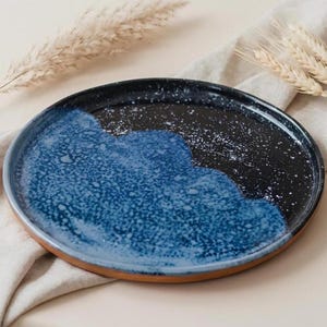 Op de afbeelding: Een rond keramisch bord met een glanzend zwart oppervlak met een gespikkelde witte afwerking en een groot, getextureerd blauw glazuurgedeelte aan één kant. Het bord heeft een zichtbare terracotta-kleurige rand aan de onderkant.