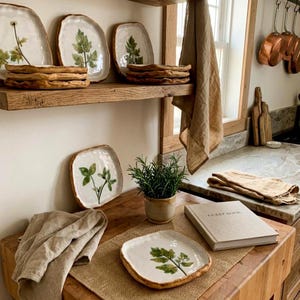 May include: Handmade ceramic plates with leaf designs. The plates are white with a brown rim and green leaf illustrations. The plates are stacked on a wooden shelf and table, with a potted plant and a guest book. The plates are ideal for serving food.