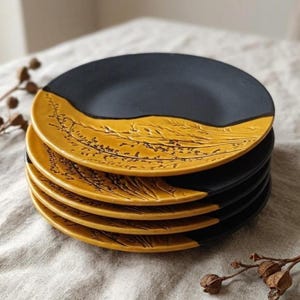 May include: A stack of six ceramic plates. The top half of each plate is a matte black, while the bottom half is a mustard yellow with a detailed botanical design. The plates are stacked on a neutral-colored surface.