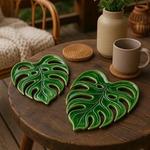 May include: Two glossy, green ceramic monstera leaf-shaped coasters sit on a dark wooden table. The coasters have a detailed vein pattern and cut-out sections. A brown mug and a woven coaster are also on the table.