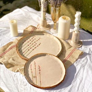May include: A still-life arrangement featuring two ceramic plates with floral designs, surrounded by candles and a glass of lavender. The plates have a light beige color with brown rims. The candles are white and beige, and the lavender is purple.