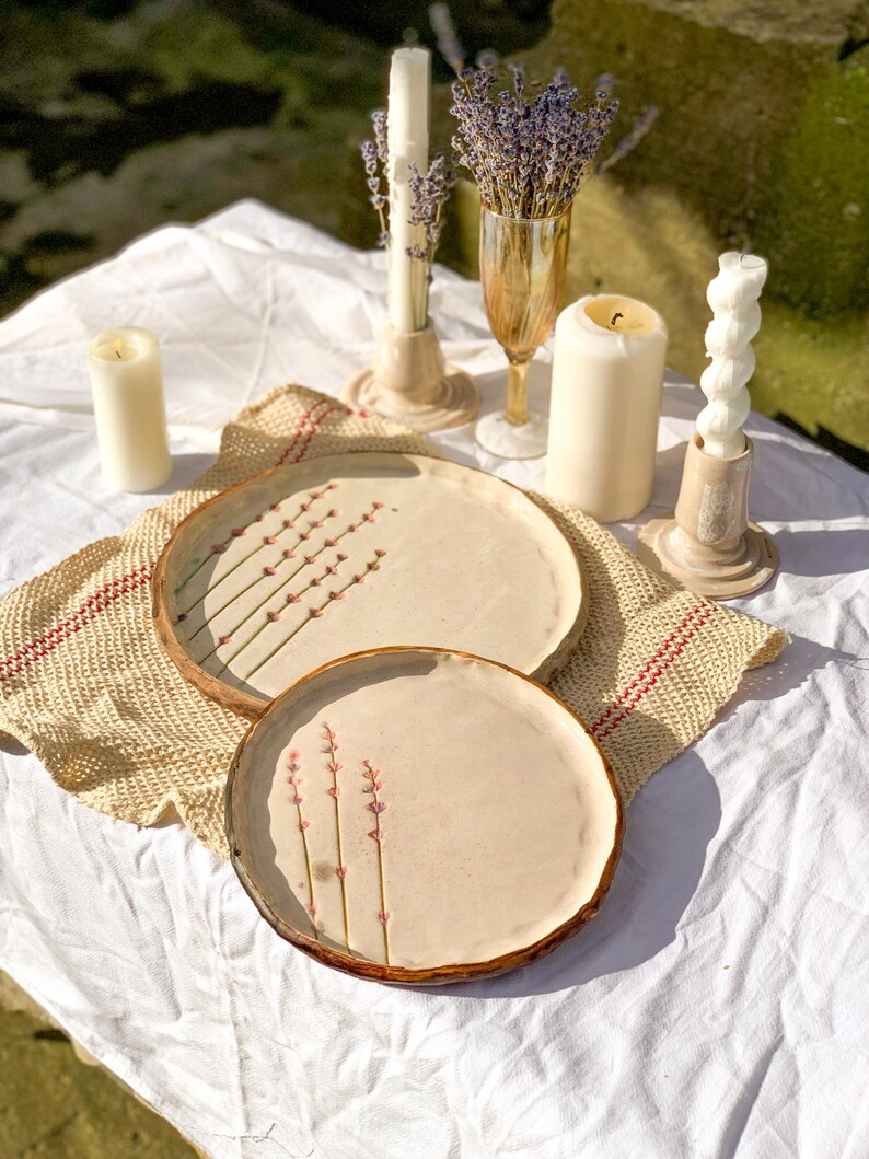 May include: A still-life arrangement featuring two ceramic plates with floral designs, surrounded by candles and a glass of lavender. The plates have a light beige color with brown rims. The candles are white and beige, and the lavender is purple.