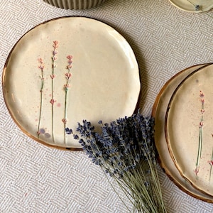 May include: Cream-colored ceramic plates with a hand-painted lavender design and a brown rim. The plates are round and have a rustic, artisanal feel. A bundle of dried lavender is placed near the plates.