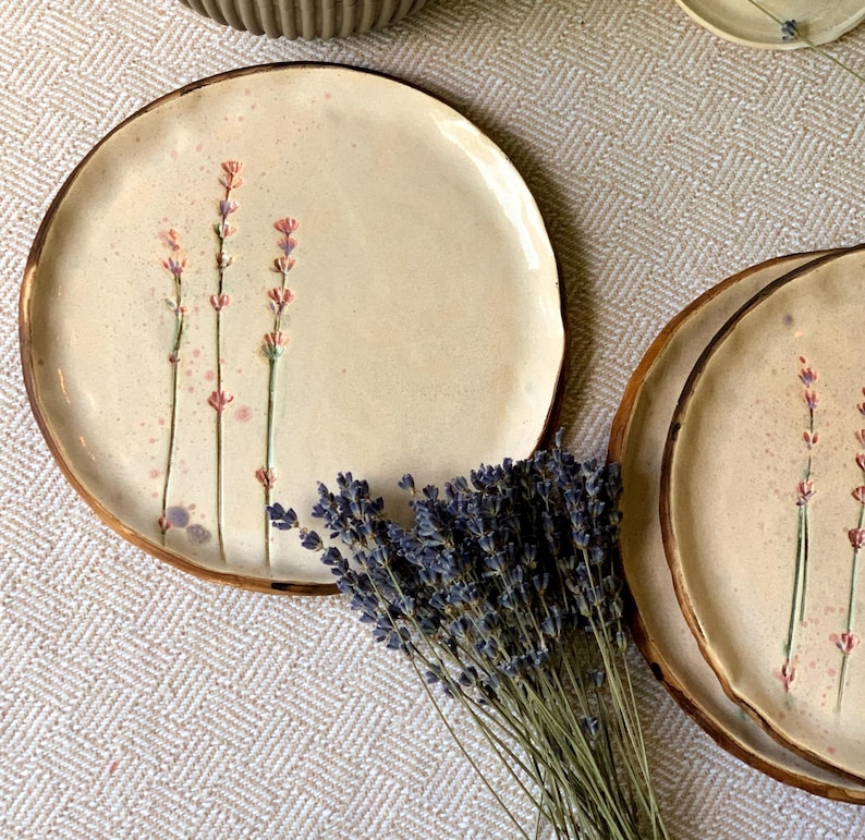 May include: Cream-colored ceramic plates with a hand-painted lavender design and a brown rim. The plates are round and have a rustic, artisanal feel. A bundle of dried lavender is placed near the plates.