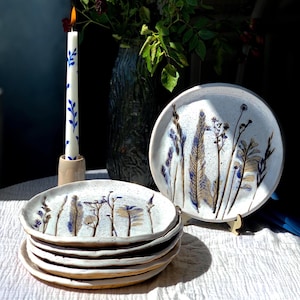 May include: A stack of white ceramic plates with a hand-painted botanical design in blue and brown. A matching plate is displayed on a wooden stand. A lit candle with blue floral accents and a vase with flowers are in the background.