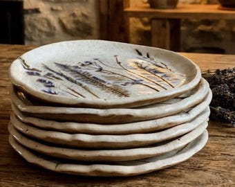 May include: A stack of six round ceramic plates with an off-white speckled glaze. Each plate features a hand-painted botanical design in shades of blue and brown, depicting delicate grasses and wildflowers with wavy, uneven edges.