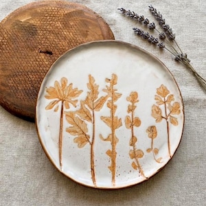 Op de afbeelding: Een witte keramische plaat met een botanisch ontwerp in bruintinten. De plaat heeft een verhoogd bloemmotief en een bruine rand. Een houten serveerplank en lavendeltakjes staan op de achtergrond.