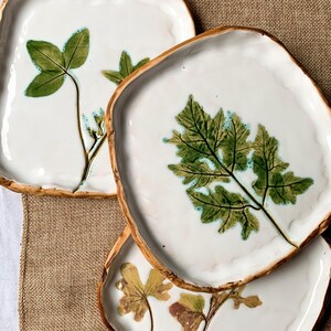 May include: White ceramic plates with a leaf design. The plates have a square shape with a wavy edge and a brown rim. The leaves are green and brown, and the plates are arranged on a beige surface.
