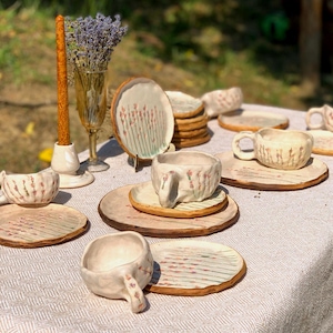 May include: A collection of handmade ceramic tableware, including cups, saucers, and plates, arranged on a white tablecloth. The pieces feature a rustic, textured finish with delicate floral patterns in shades of pink and brown. A candle and a vase of lavender complete the scene.
