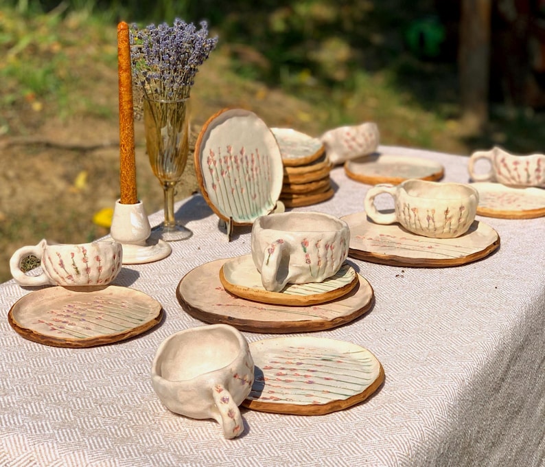 May include: A collection of handmade ceramic tableware, including cups, saucers, and plates, arranged on a white tablecloth. The pieces feature a rustic, textured finish with delicate floral patterns in shades of pink and brown. A candle and a vase of lavender complete the scene.