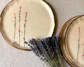 May include: Cream-colored ceramic plates with a brown rim and a lavender floral design. The plates have a rustic, handmade appearance. A bundle of dried lavender is placed near the plates, adding a natural element to the composition.