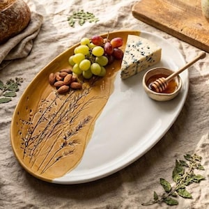 Puede incluir: Una bandeja de servicio ovalada de cerámica con un diseño bicolor, que presenta una sección blanca y otra de color amarillo mostaza con un patrón botánico de trigo en relieve. Contiene uvas verdes, almendras, queso azul y miel.