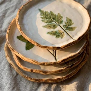 May include: A stack of handmade ceramic plates with a white glaze and a natural, organic edge. Each plate features a detailed green fern leaf design. The plates have a rustic, earthy aesthetic, perfect for serving food.