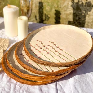 Op de afbeelding: Een stapel van vier keramische borden met een crèmekleurig oppervlak en een bruine, getextureerde rand. Elk bord is versierd met een delicaat bloemmotief van roze knoppen op groene stelen. De borden zijn op een wit tafelkleed geplaatst.