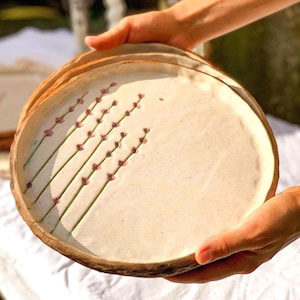 May include: A round, off-white ceramic plate with a textured, speckled surface. The plate features a hand-painted design of several lavender sprigs with green stems and small purple flowers arranged diagonally across the left side of the surface.