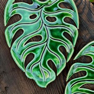 May include: Two glossy, green ceramic leaf-shaped coasters with a monstera plant design. The coasters have a dark green outline and are set on a dark wooden surface. The design features several oval-shaped cutouts.