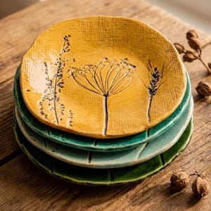 May include: A stack of three round ceramic plates with textured surfaces. The top plate is mustard yellow with black botanical illustrations of wildflowers and wheat. The middle plate is light teal, and the bottom plate is dark forest green.