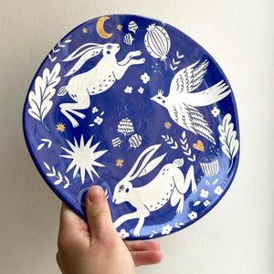 Puede incluir: Un plato de cerámica redondo con un esmalte azul profundo que presenta ilustraciones blancas de dos liebres corriendo, un pájaro en vuelo, hojas, flores y vainas. El diseño incluye pequeños detalles dorados en forma de luna creciente, estrellas y un corazón.