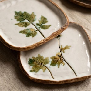 May include: Two square ceramic plates with a white surface and a brown, textured rim. Each plate features a green and yellow leaf design. The plates are ideal for serving appetizers or small dishes.