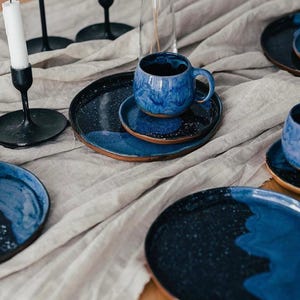 Può includere: Una tavola apparecchiata con tazze, piattini e piatti in ceramica blu con un design maculato. Le tazze hanno un manico e poggiano su piattini abbinati. I piatti sono blu scuro con un motivo maculato.