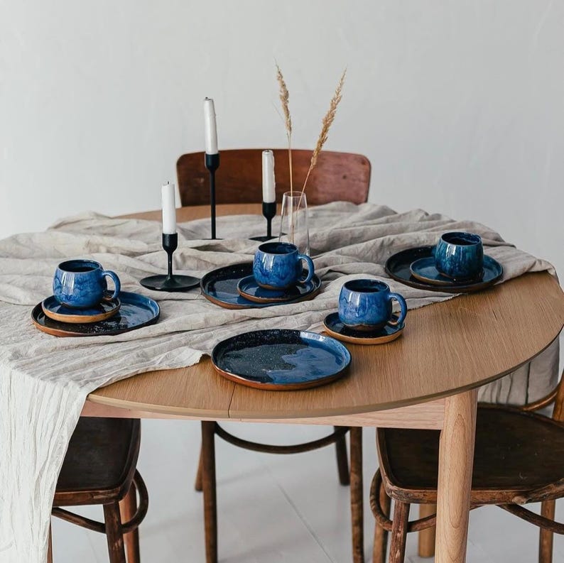 May include: A wooden table set with blue ceramic mugs, saucers, and plates. The mugs and plates have a speckled glaze. Black candle holders with white candles and a vase with dried grass are also on the table. The table is covered with a beige linen tablecloth.