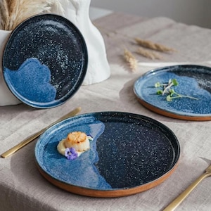 May include: A set of three ceramic plates with a speckled navy blue and light blue design. The plates have a unique wave-like pattern and a brown rim. One plate is garnished with food and a gold knife.